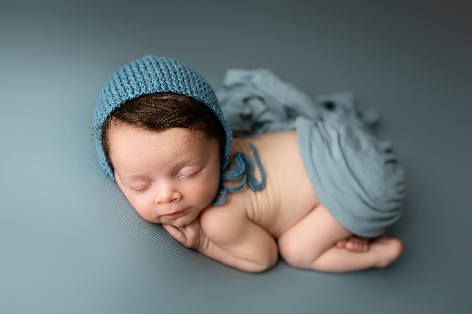 Infant boy posed on blue wearing a blue bonnet.