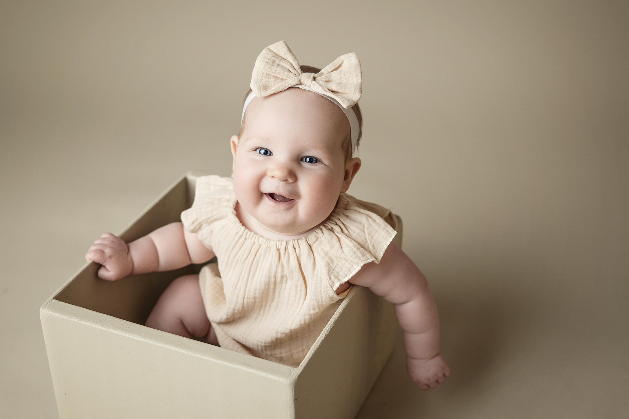 Smiling baby girl posed in a cream colored box wearing a matching romper and bow headband.
